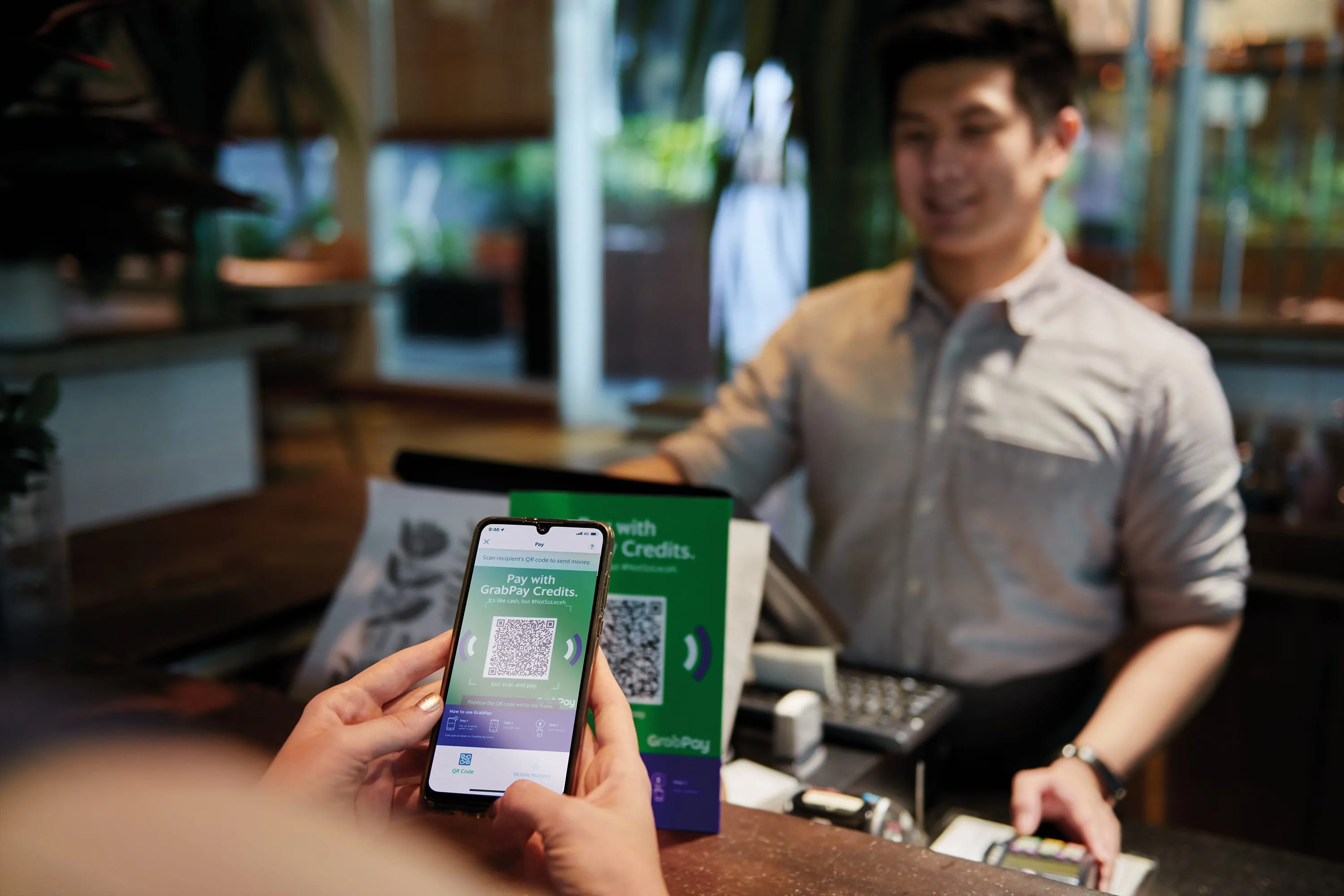 In the foreground, a customer scans a GrabPay QR code. In the background is a service staff with a smile, in a cafe.